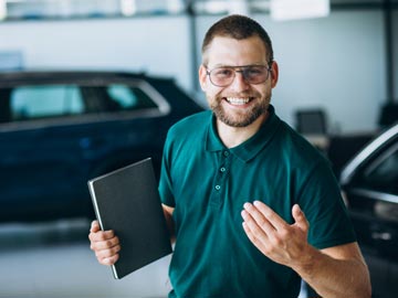 Ein Mann in einem grünen Polo-Shirt hält ein Notizbuch in der Hand und gestikuliert, während er in einem Autohaus steht, umgeben von Fahrzeugen.