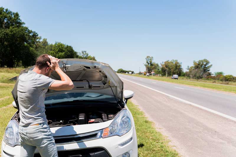 Ein besorgter Mann steht neben seinem weißen Auto mit offener Motorhaube an einem sonnigen Straßenrand und sieht verwirrt und besorgt wegen Autoproblemen aus.