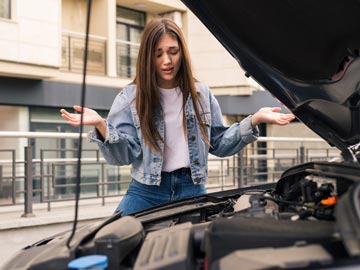 Eine junge Frau in einer Jeansjacke steht neben einer offenen Motorhaube, sieht verwirrt aus und gestikuliert mit ihren Händen.